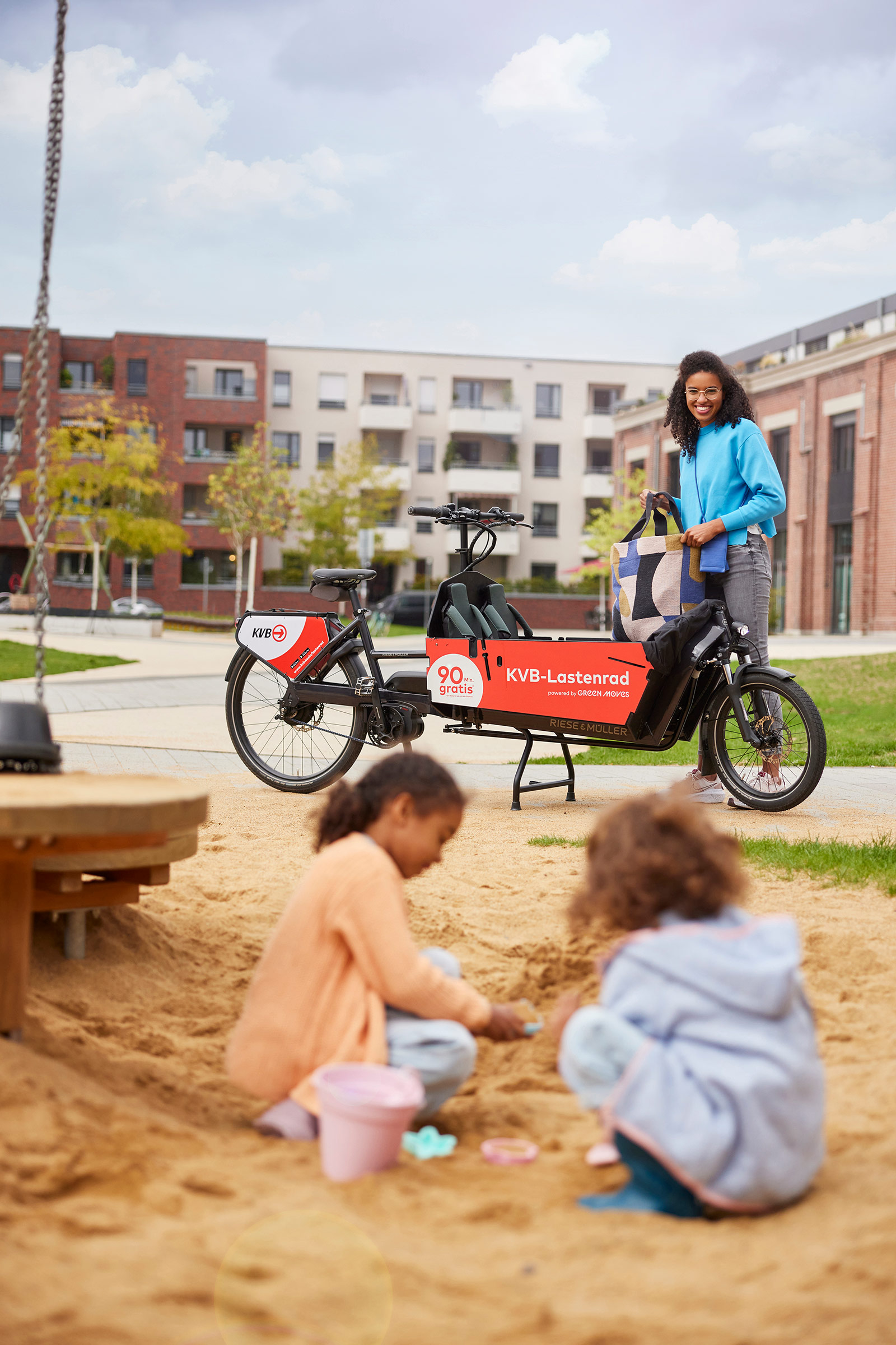 Frau neben einem KBV-Lastenrad mit Kindern auf einem Spielplatz im Wohngebiet